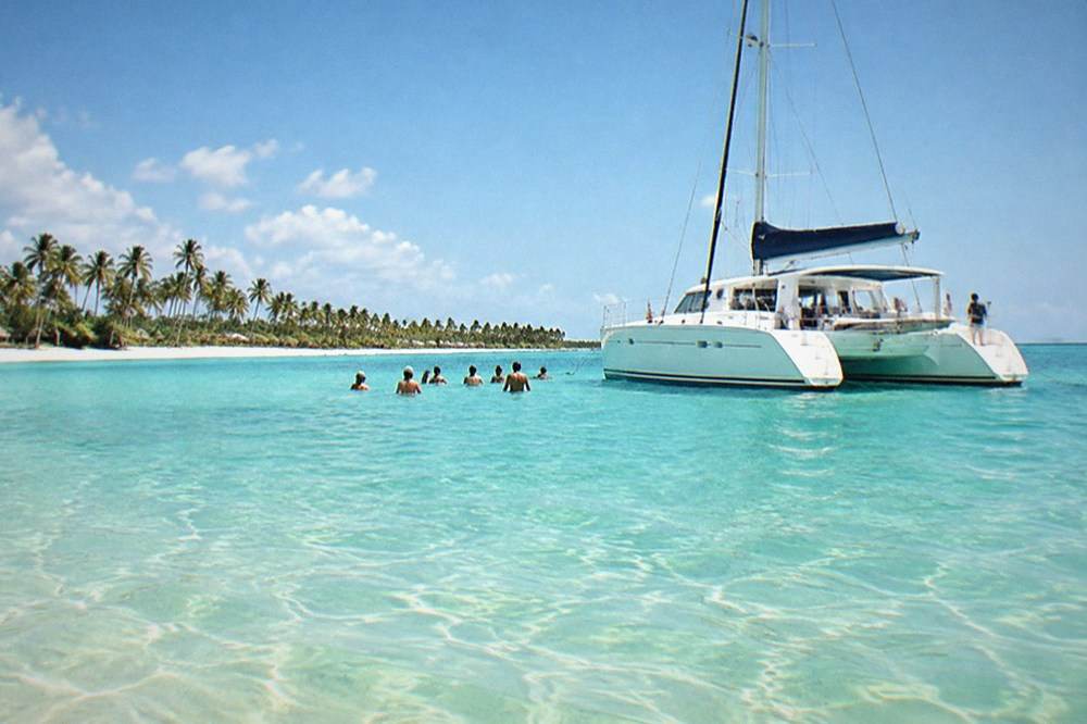 Catamaran with passengers aboard floating over clear shallow turquoise water near a sandbar
