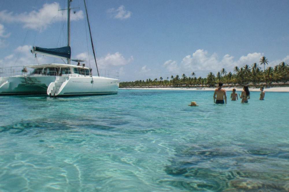 A catamaran anchored over shallow turquoise water with guests standing on a sandbar