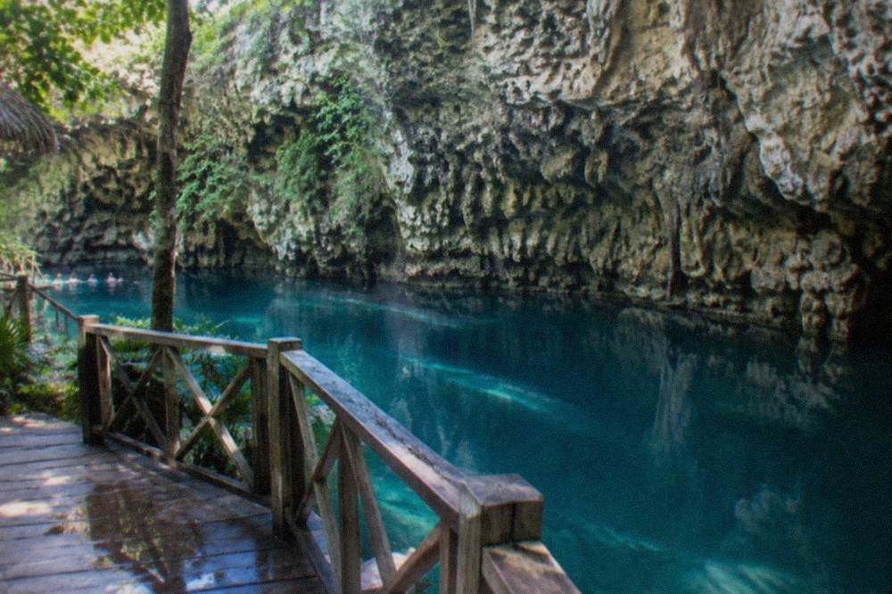 Clear blue-green cenote water seen from a wooden platform beneath a tall rock cliff.