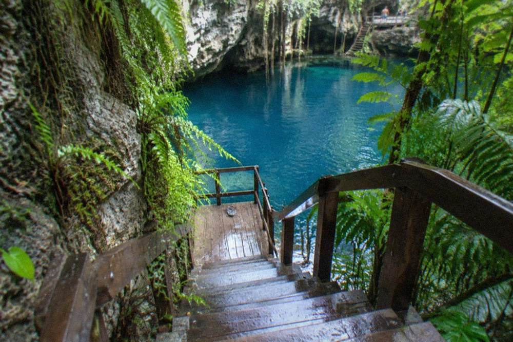 Wooden staircase and platform descending along a rock face toward still blue water in a jungle cenote.