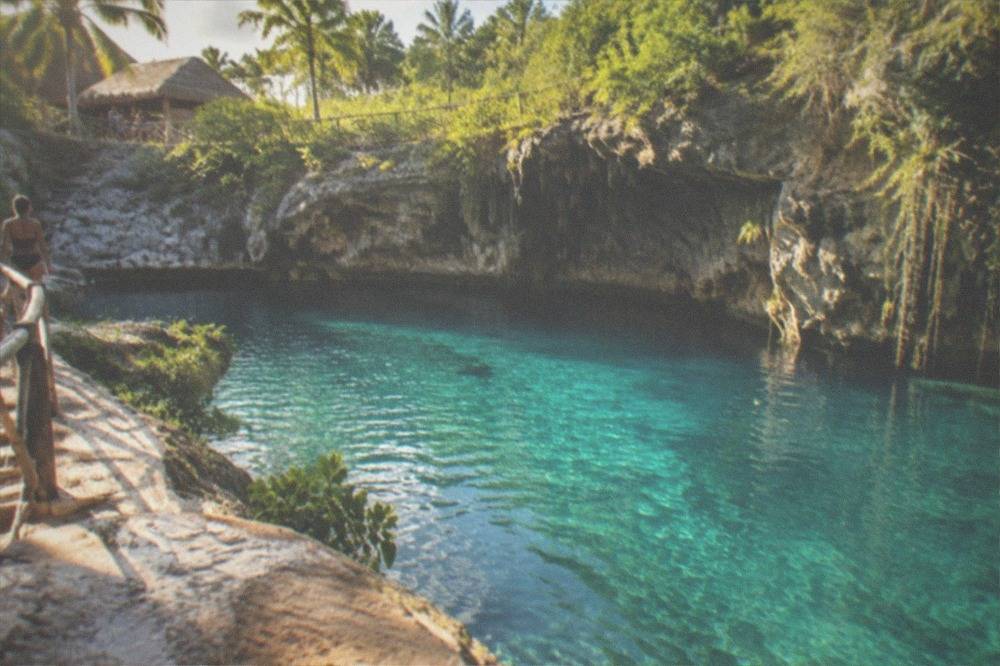 Sunlit turquoise water filling a rock basin at the base of a tall cliff, dense green plants on the ledges above.