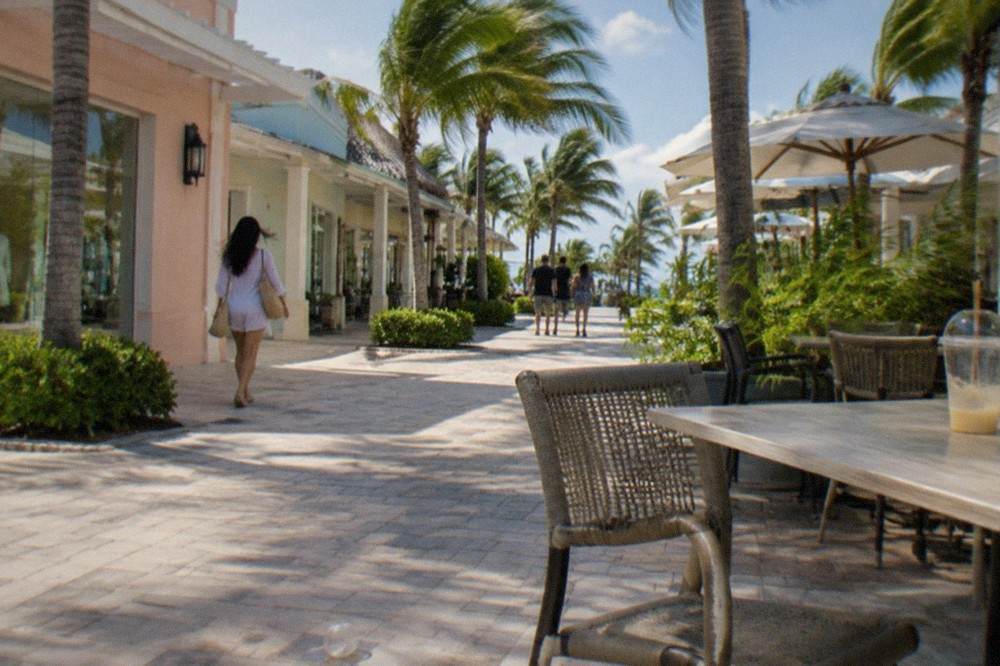 Busy outdoor mall corridor with palm trees, shops and cafes on both sides, set against a bright blue sky