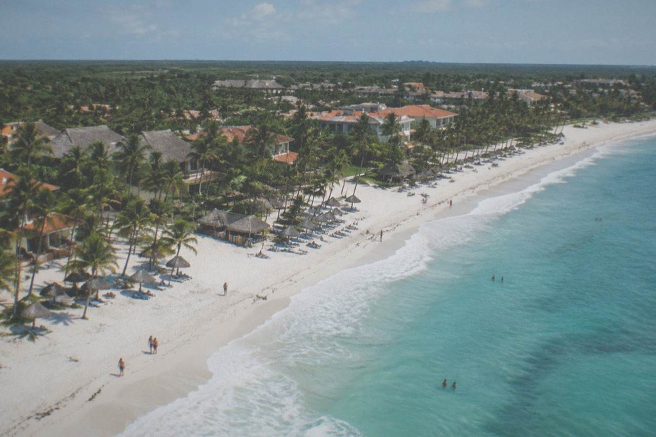 Aerial view of a resort-lined beach and a cluster of shops and restaurants in the distance, showing the spread-out layout of Punta Cana without a central downtown
