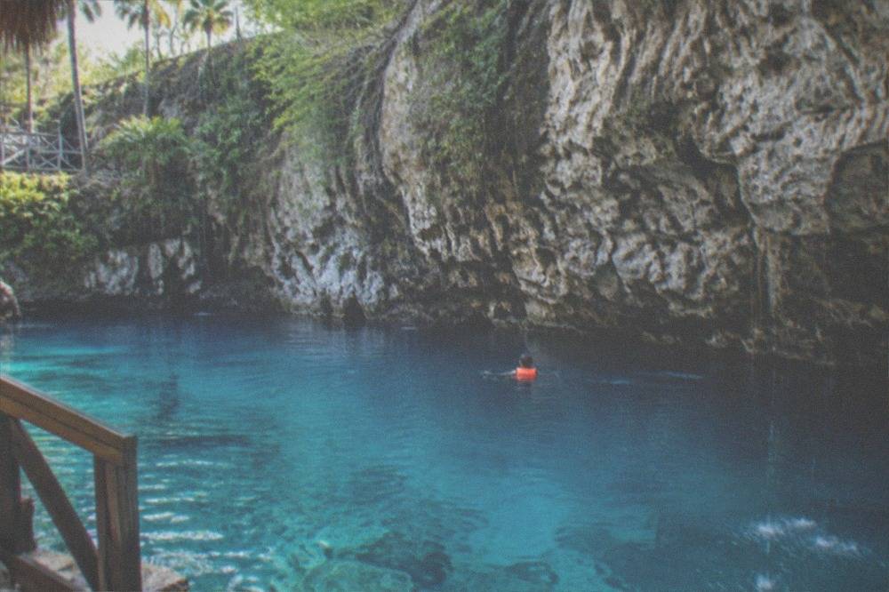 A swimmer in a life jacket floating in clear blue cenote water beside a rock wall