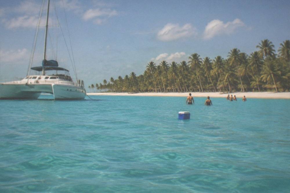 Catamaran moored offshore with passengers wading in shallow turquoise water near a palm-lined beach