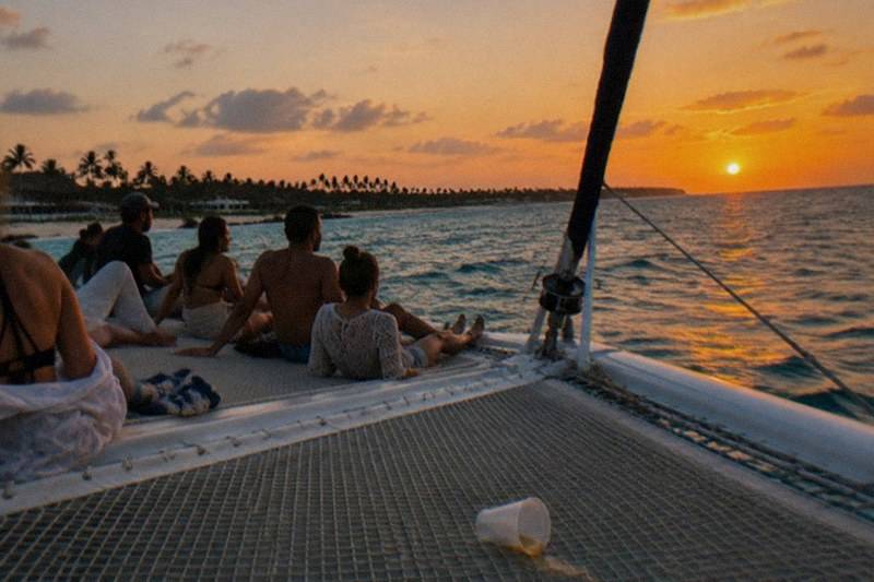Passengers lean on the rails of a catamaran as it sails the Punta Cana coast at sunset, the sky streaked orange over open water.