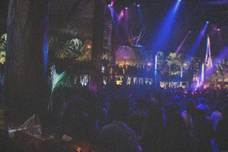 Spotlights sweep across a packed theater crowd at a Punta Cana show venue, an aerial rig and mirrored backdrop catching the colored beams.