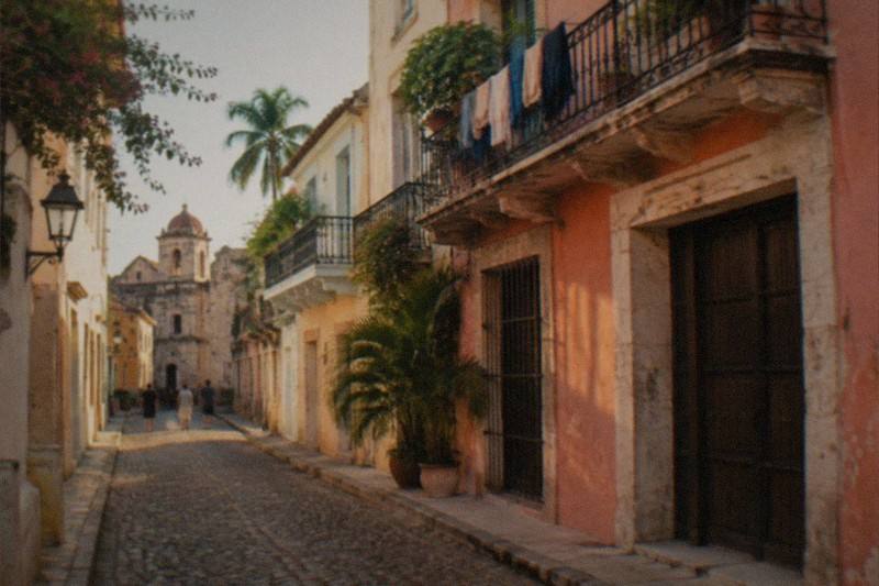 A cobblestone colonial street lined with pale stone buildings and wrought-iron balconies, warm afternoon light falling across the facades.