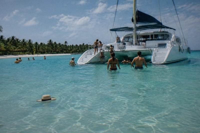 A catamaran floats in waist-deep turquoise water beside a sandbar, passengers wading and laughing under a bright midday sky.