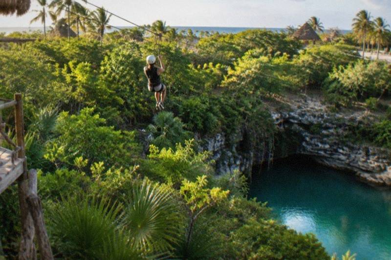 A rider in a harness crosses a long zipline above dense jungle canopy, the blue water of a cenote far below the cable.