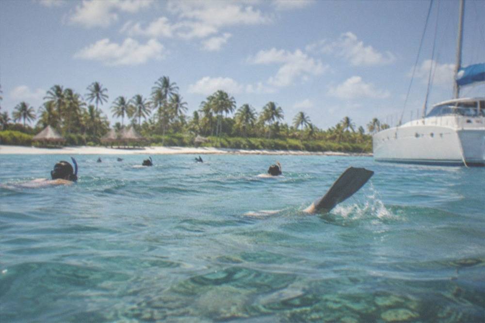 Passengers in masks and fins snorkeling over a shallow reef from a moored catamaran