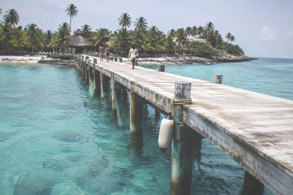 Long wooden pier extending over clear shallow water toward a palm-lined island beach