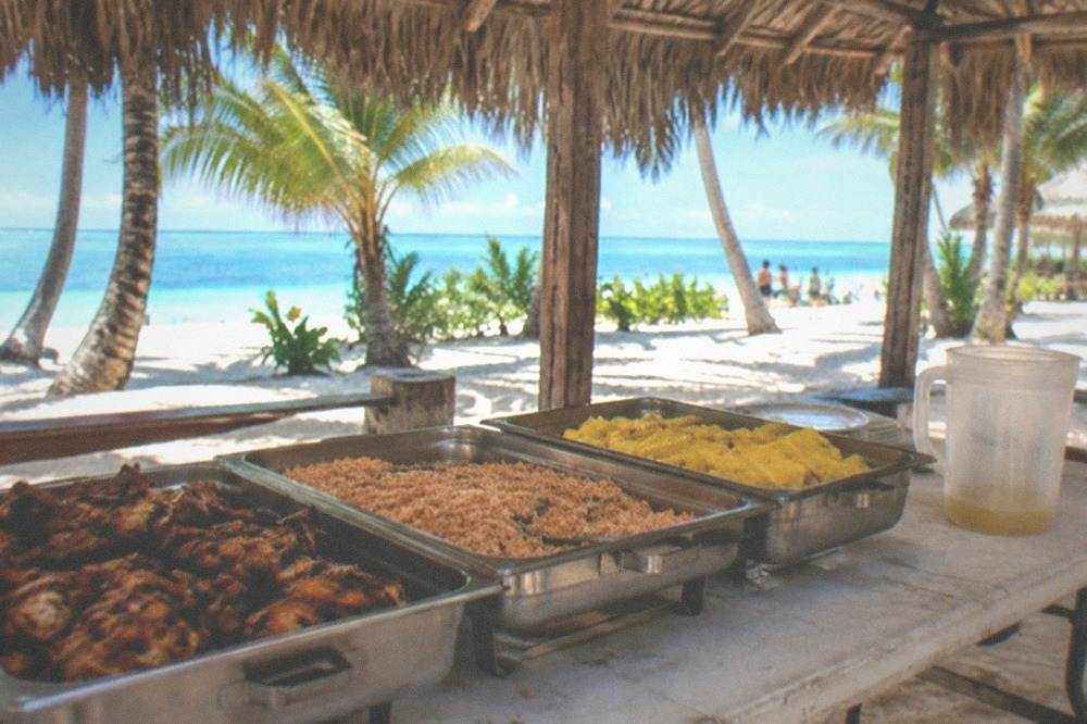 An open-air buffet table with grilled food and tropical fruit under a palm-thatch roof